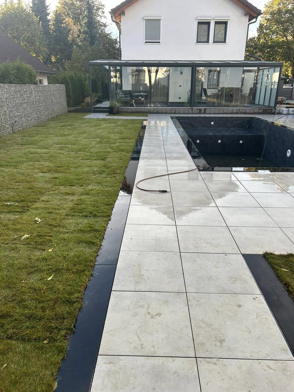 Poolanlage mit hellen Feinsteinzeugplatten, Rollrasen und Gabionenwand vor einem Haus mit Glasanbau